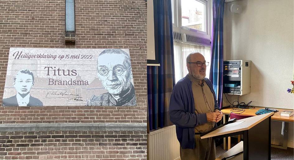 A man stands in an office setting beside a memorial plaque of Titus Brandsma on a brick wall.