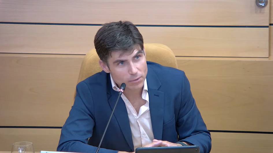 A man in a dark suit and white shirt sits at a desk with a microphone, looking pensive.