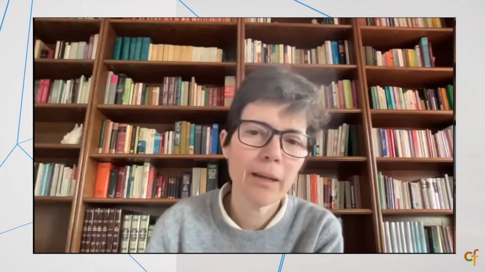 A person with glasses in a grey sweater is speaking in front of a bookshelf filled with various books.