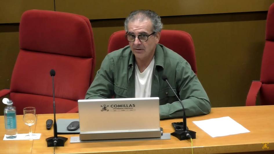 A man sitting at a desk with a laptop in a conference room with a label 'Comillas' on the desk.