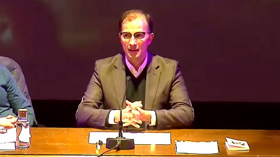 A man wearing glasses is seated at a table with microphones during a conference.