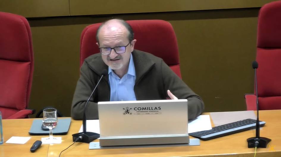 A man in a dark jacket and glasses sits at a table speaking, with a laptop in front of him and a sign reading 'Comillas' on it in a lecture hall.