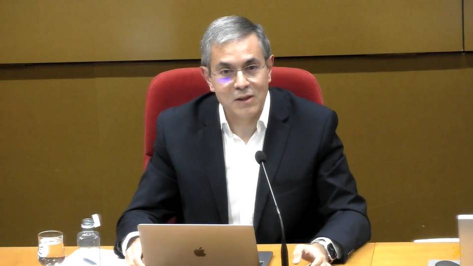 A man in a white shirt and black jacket sitting at a desk with a laptop, speaking at a conference