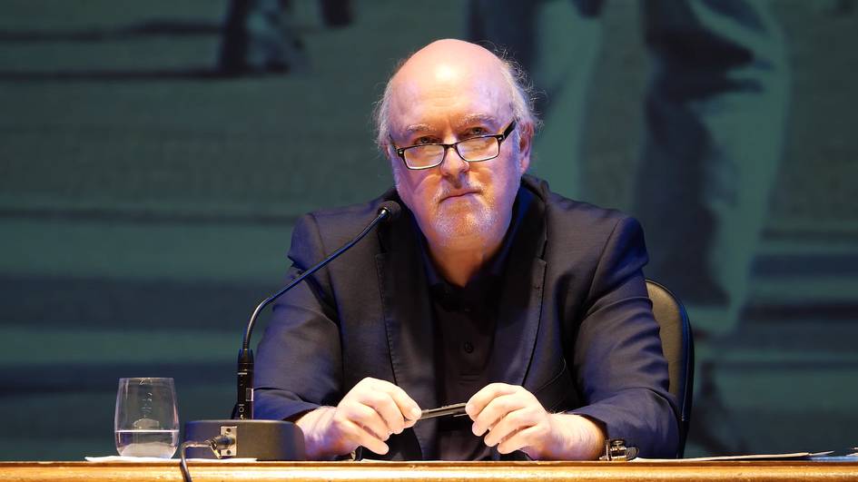 A man in glasses sits at a desk with a microphone, appearing to be engaged in a discussion or presentation at a public event.