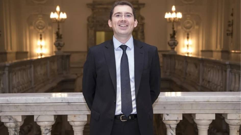 Un hombre sonriente vestido de traje se encuentra de pie en un vestíbulo elegante con decoración clásica.