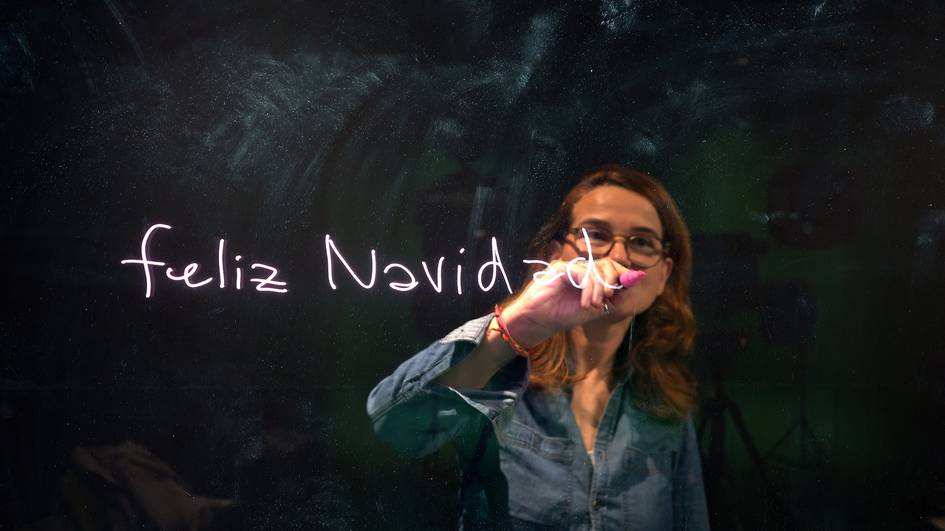 Una mujer escribe 'feliz Navidad' en un cristal con una luz brillante.