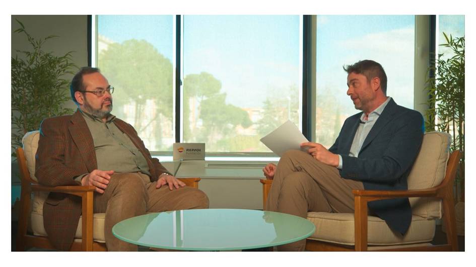 Two men seated in armchairs having a discussion in a room with large windows and a view of trees.