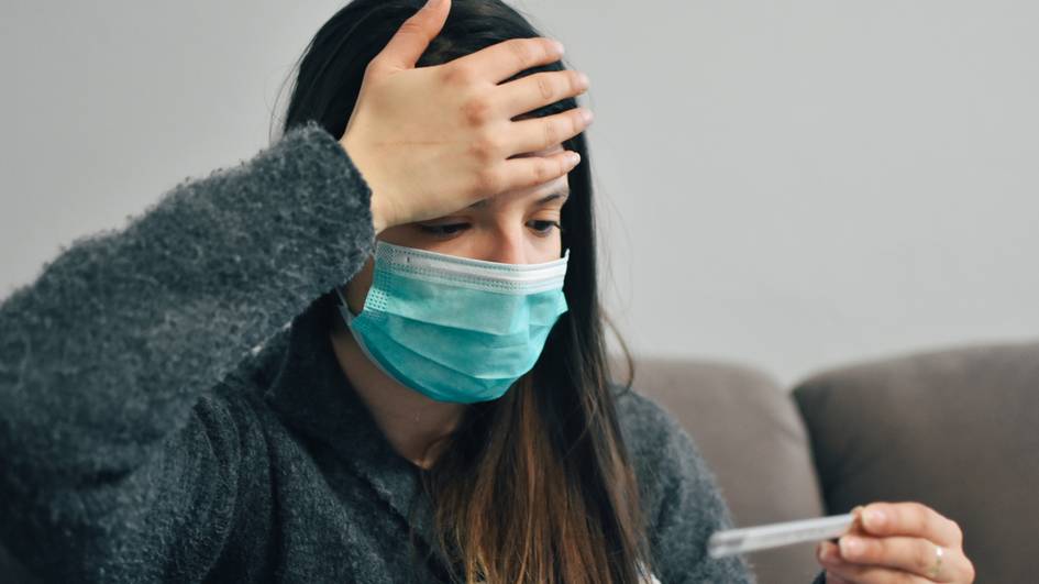 Una mujer con mascarilla sostiene un termómetro con expresión de preocupación.