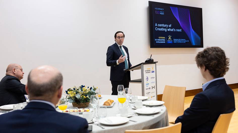 Un hombre habla en un evento mientras otras personas lo escuchan desde una mesa decorada con comida.