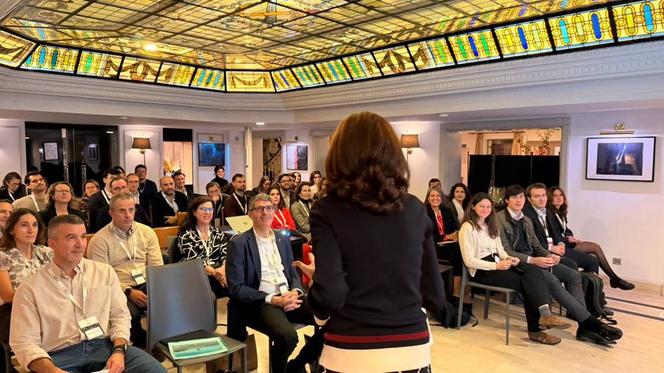Una conferencia con un gran público en sala iluminada por un techo de vidrio colorido.