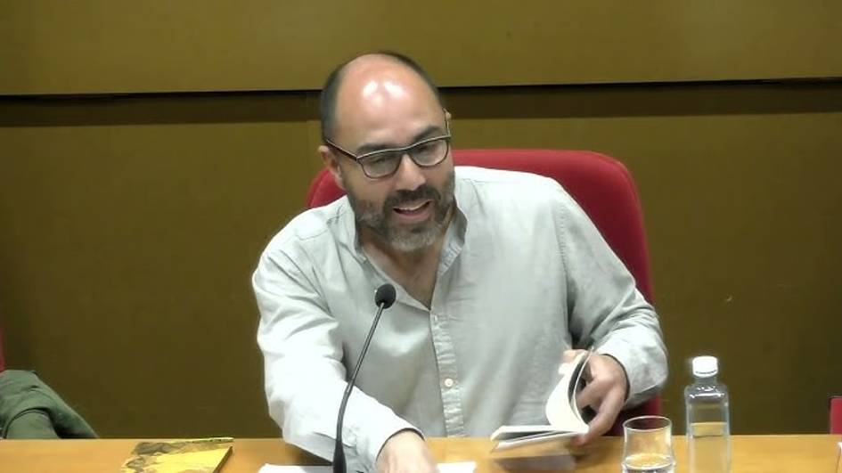 Un hombre con gafas y barba, leyendo un libro en un evento público con un micrófono y agua al frente.