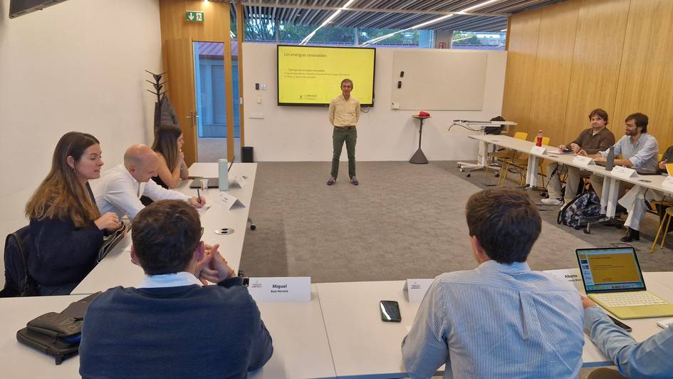 Una reunión en un entorno moderno donde un presentador está hablando frente a un grupo sentado alrededor de una mesa.