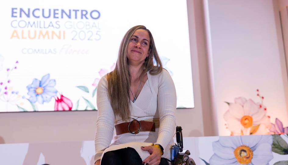 Una mujer sonriente en una conferencia, sentada en una silla de ruedas, frente a un fondo decorativo.