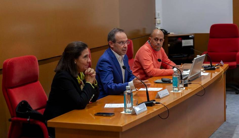 Three professionals, two men and one woman, are seated at a panel in a conference room.