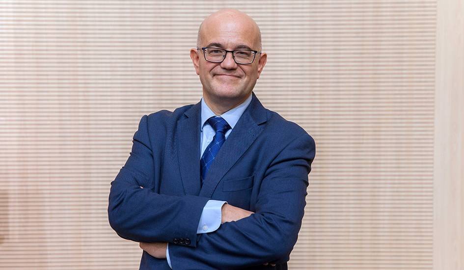 A man in a blue suit and glasses standing confidently with crossed arms in an office setting.