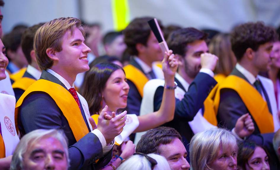 A group of young graduates in orange stoles celebrating at a graduation ceremony, one young man in the foreground is holding a diploma.