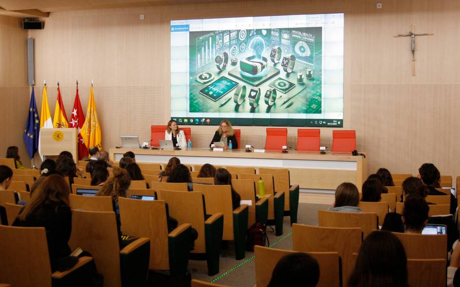 A conference room with attendees watching speakers at the front and an on-screen digital technology graphic.