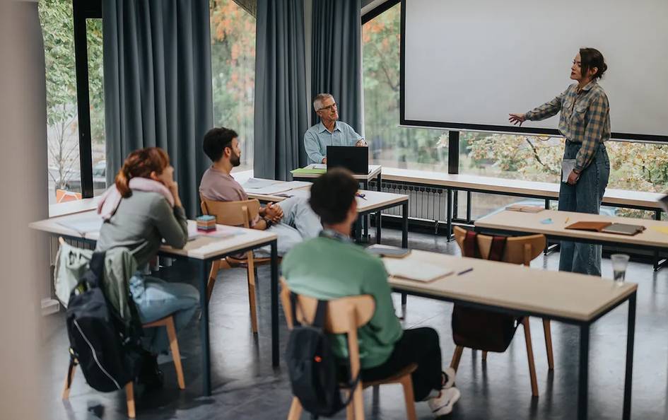 Una mujer presenta en una clase a un grupo de estudiantes atentos en un aula moderna.