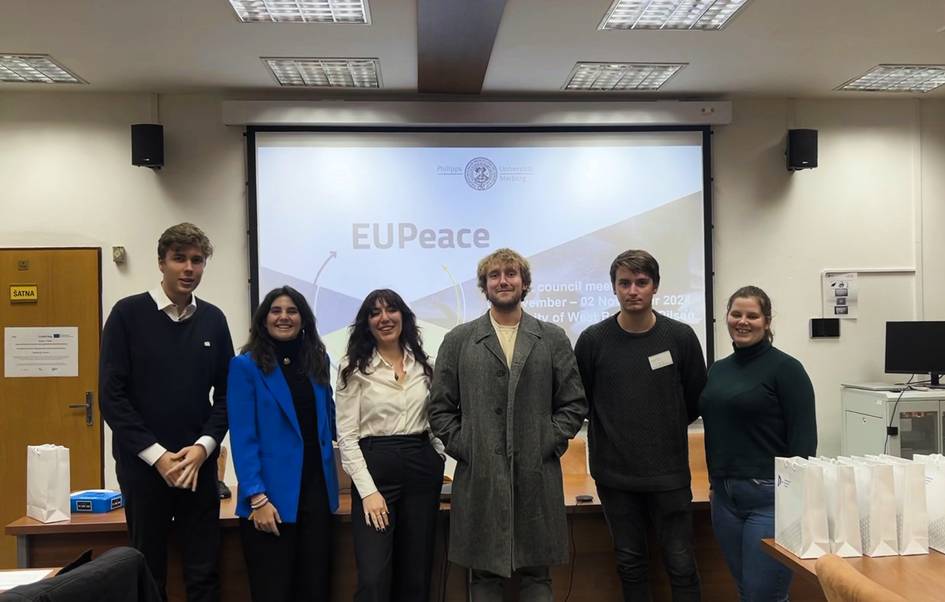 Grupo de seis personas jóvenes sonriendo en una sala de conferencias con una presentación de EUPeace en la pantalla.