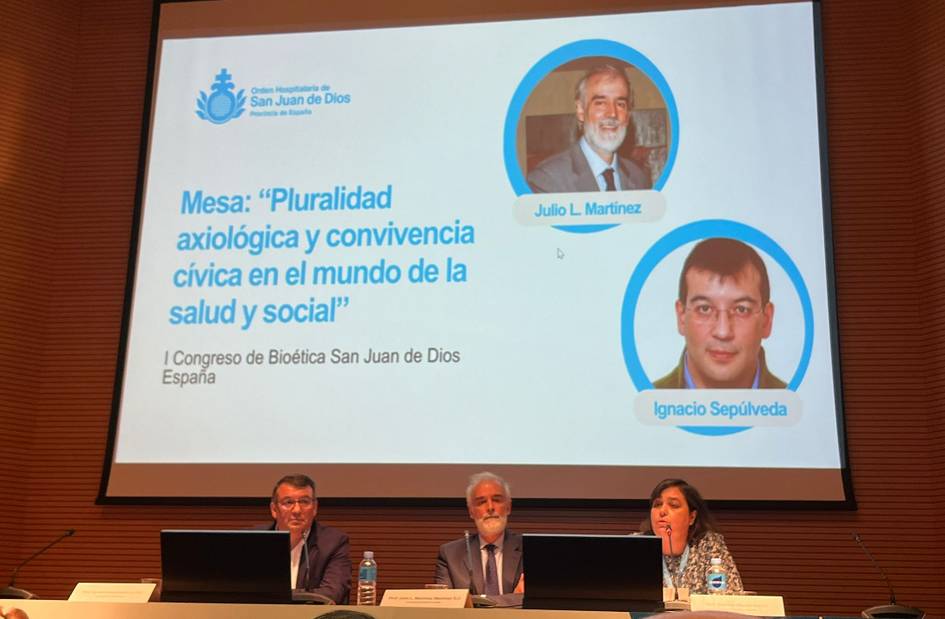 Three professionals sitting at a panel discussion table during a conference on bioethics, with a presentation slide visible in the background.