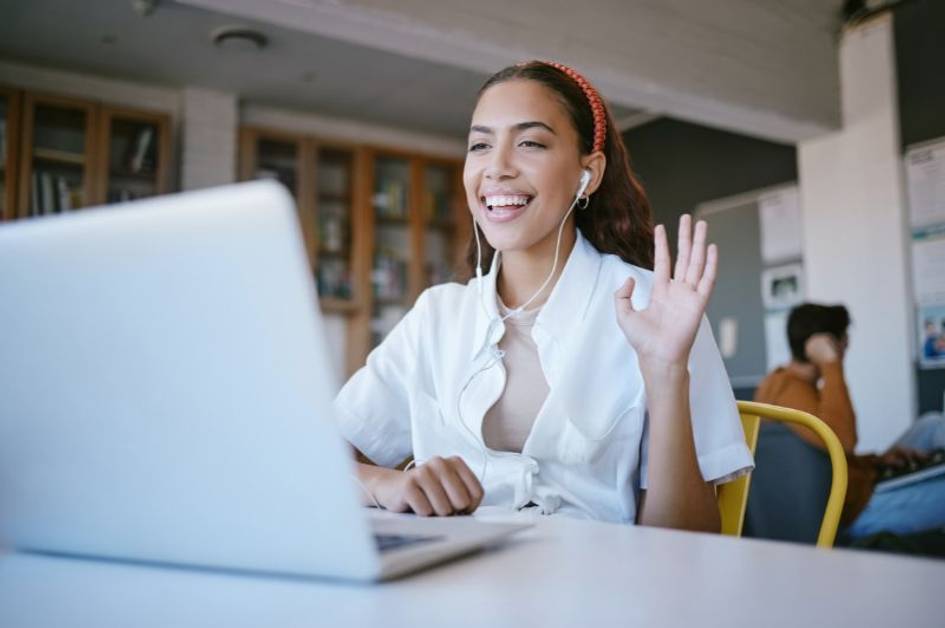 A smiling young woman with headphones waves at her laptop screen, likely engaging in a video call, in a brightly lit indoor setting.