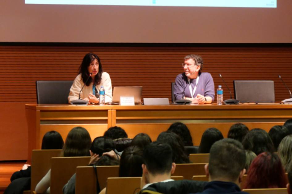 Una conferencia en un auditorio con dos ponentes en el escenario y un público atento.