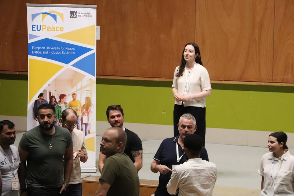 Un grupo de personas se agrupa alrededor de una pancarta que promueve la Universidad Europea para la Paz, mientras una mujer se encuentra de pie detrás de ellos.