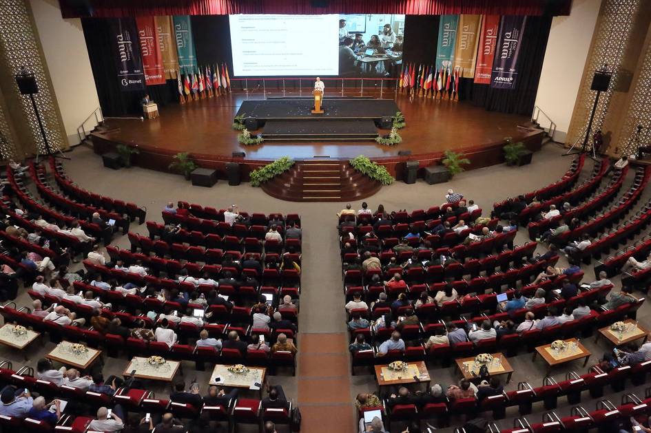 Una gran sala de conferencias con un escenario y una audiencia numerosa escuchando a un ponente.