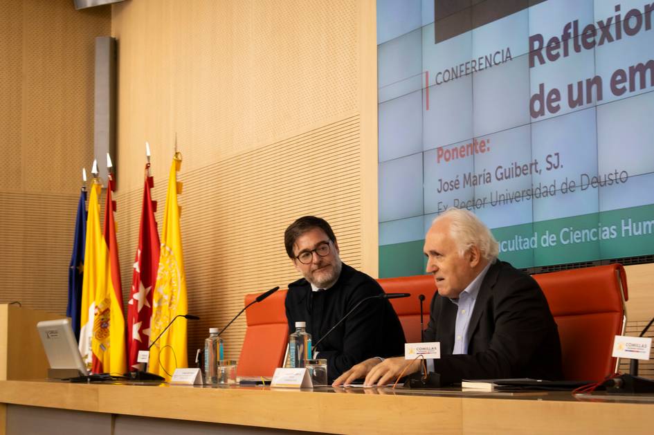 Dos hombres están sentados en un panel de discusión en una conferencia, con banderas y un proyector en el fondo.