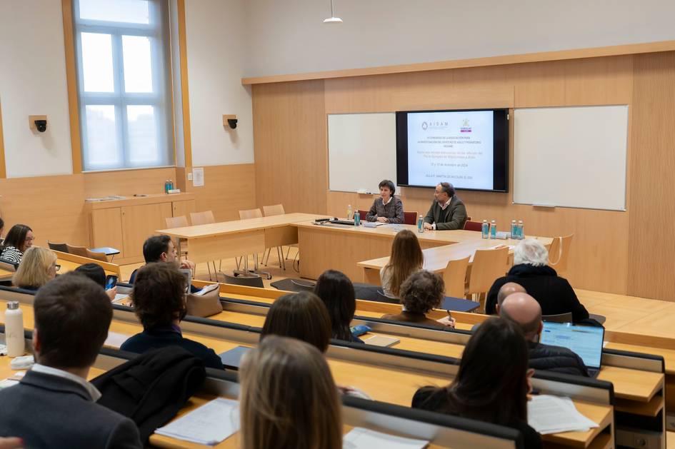 Dos personas conversando en un panel frente a una audiencia en un aula universitaria.