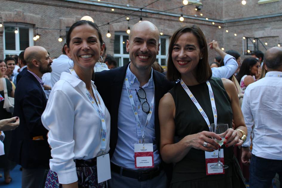 Un grupo de tres amigos sonríen en un evento social al aire libre rodeados de personas.