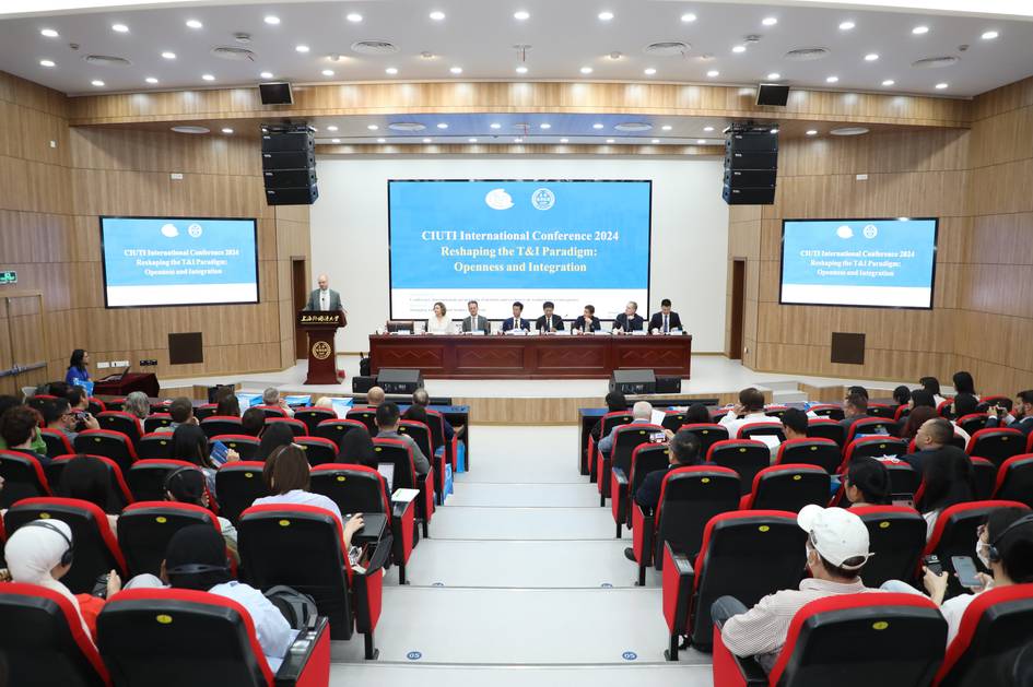 A conference symposium scene featuring multiple speakers at the front and an audience of attendees facing the podium in a modern hall.