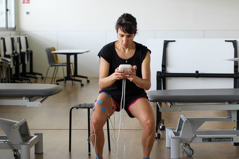 A woman sitting in a rehabilitation center using a mobile device while connected to a muscle stimulation machine.