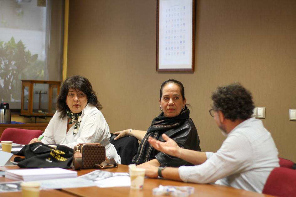 Tres personas participan en una reunión en una sala con mesa y documentos.