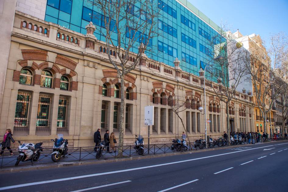 La imagen muestra un edificio histórico a la izquierda y una moderna estructura de vidrio a la derecha, con personas caminando y motocicletas estacionadas en la acera.