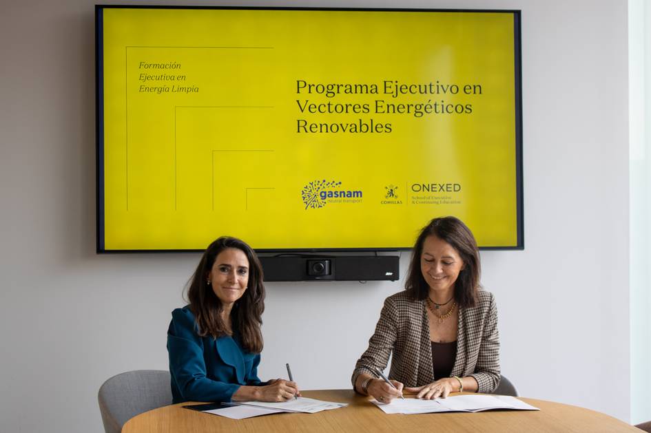 Dos mujeres se sientan en una mesa, firmando documentos frente a una pantalla que muestra información sobre un programa de energía renovable.