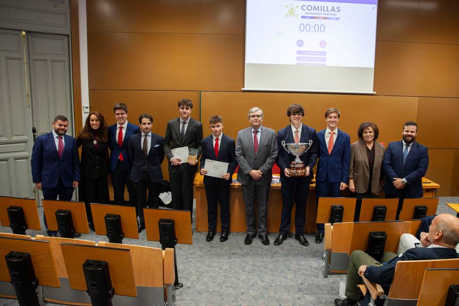 Grupo de personas vestidas formalmente, posan en un aula con diplomas y un trofeo, frente a un proyector que muestra el logo de Comillas y un cronómetro en cero.