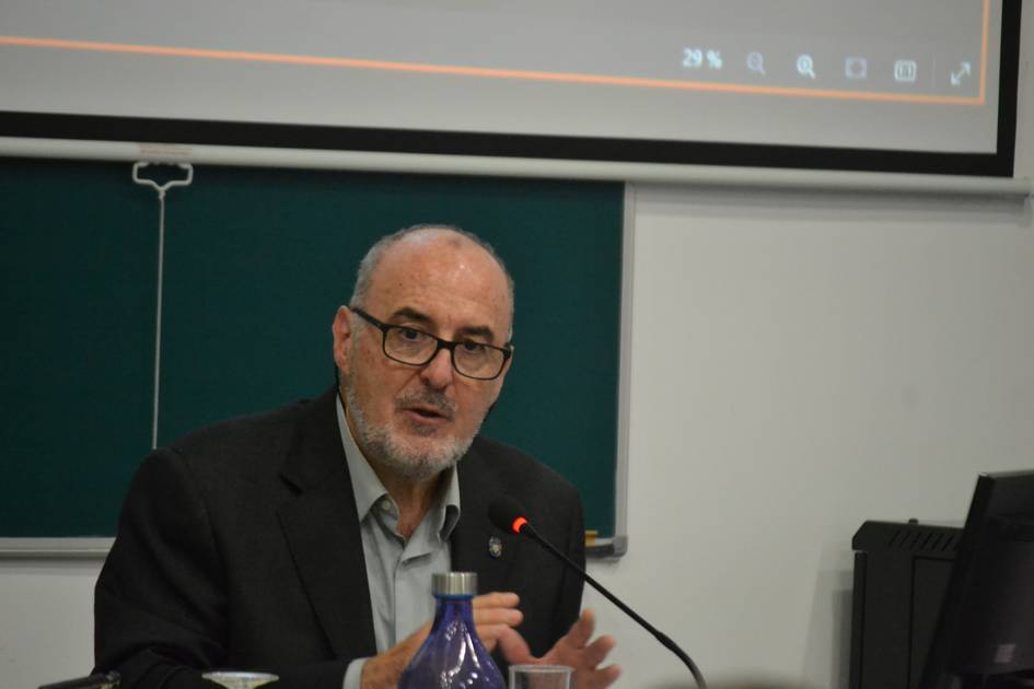 A man in a dark suit speaks at a podium in a classroom setting with a presentation screen in the background.