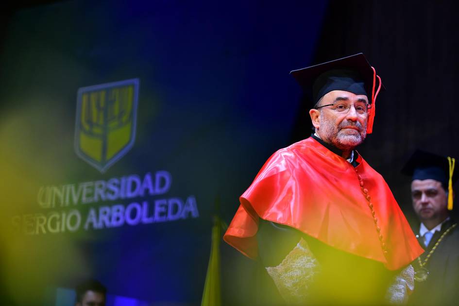 Un hombre con toga y birrete se encuentra en un escenario durante una ceremonia académica.