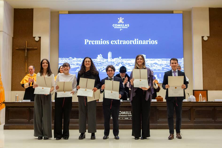 Grupo de personas recibiendo premios extraordinarios en un auditorio con un gran cartel de 'Comillas' al fondo.