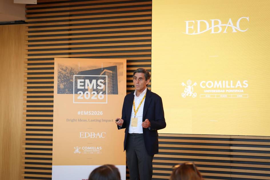 Un hombre está hablando en un evento con un cartel de fondo que muestra el logo de EMS 2026 y de la Universidad Pontificia Comillas.