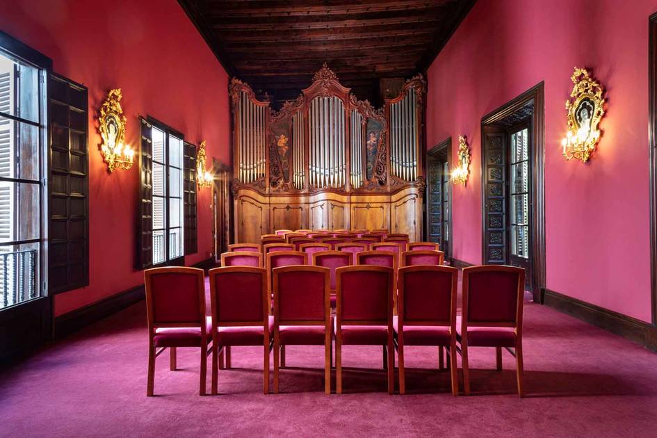 Interior de un pequeño auditorio con sillas rojas, órgano de tubos al fondo y paredes rosadas iluminadas por apliques dorados.