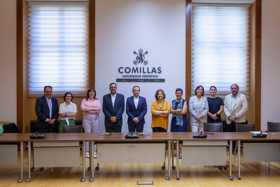 A group of nine individuals standing behind a conference table in a room with a logo of 'Comillas Pontifical University'.