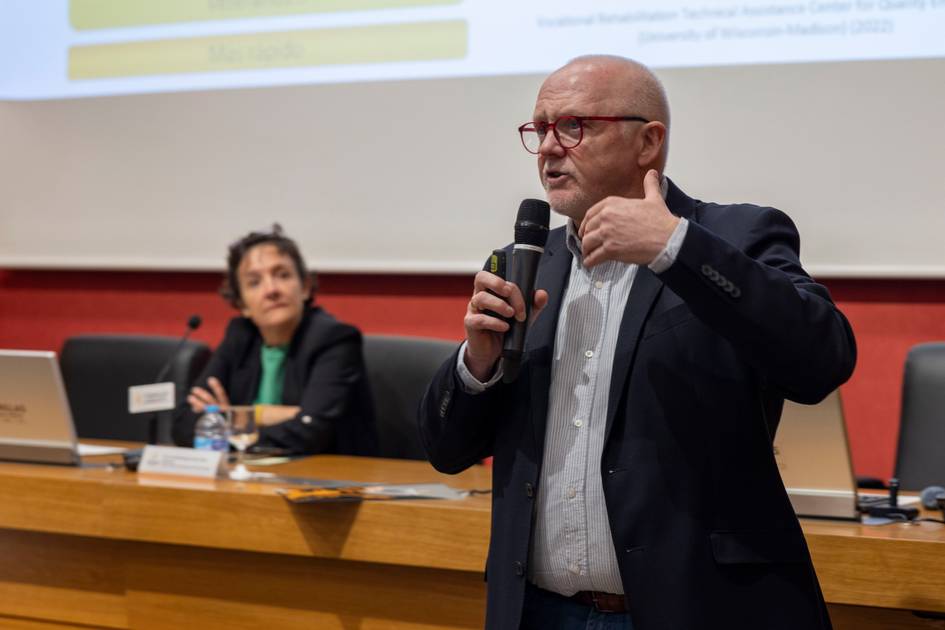 Un hombre presenta un tema en una conferencia mientras una mujer lo escucha en el fondo.