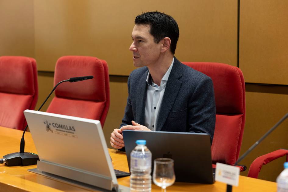 Un hombre está sentado en una mesa durante una presentación, con una computadora portátil y botellas de agua frente a él.
