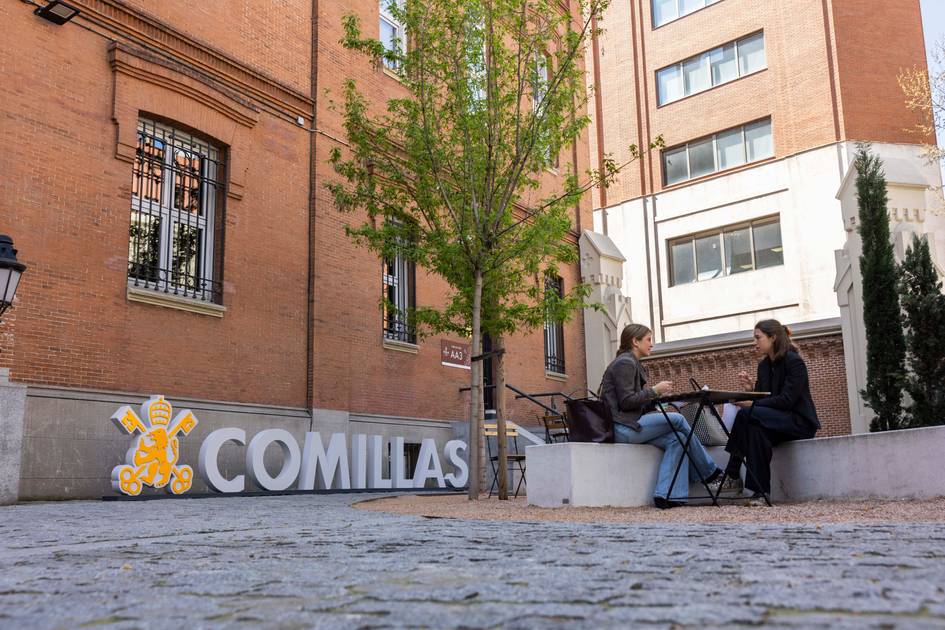 Dos personas conversan en un ambiente al aire libre frente a un edificio de ladrillo con un cartel que dice 'COMILLAS'.