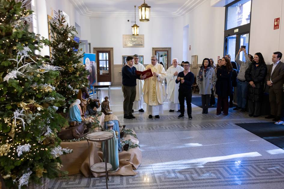 Una ceremonia navideña con un árbol decorado y figuras de Belén en un salón lleno de personas.