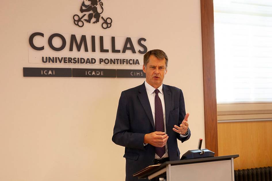 A man in a business suit is giving a speech at a podium in front of a sign that reads 'Comillas Universidad Pontificia'.
