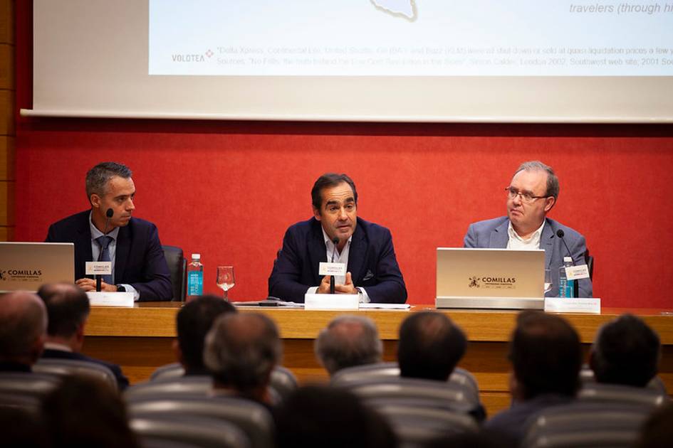 Three men sitting at a panel during a conference, addressing an audience in a lecture hall.