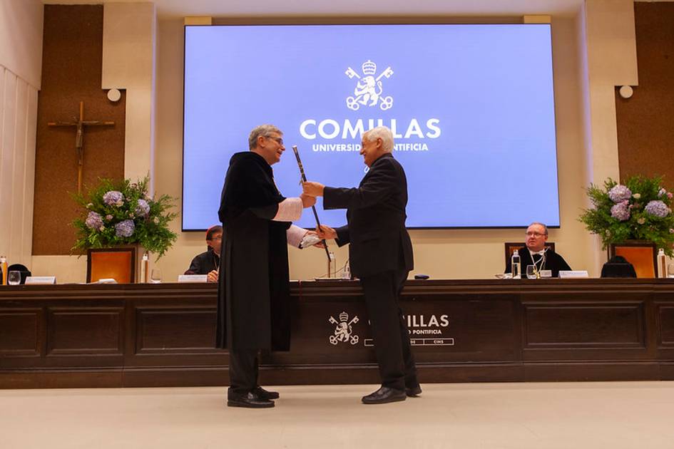 Two men shaking hands on a stage in front of a screen displaying the logo and name of Comillas Pontifical University.